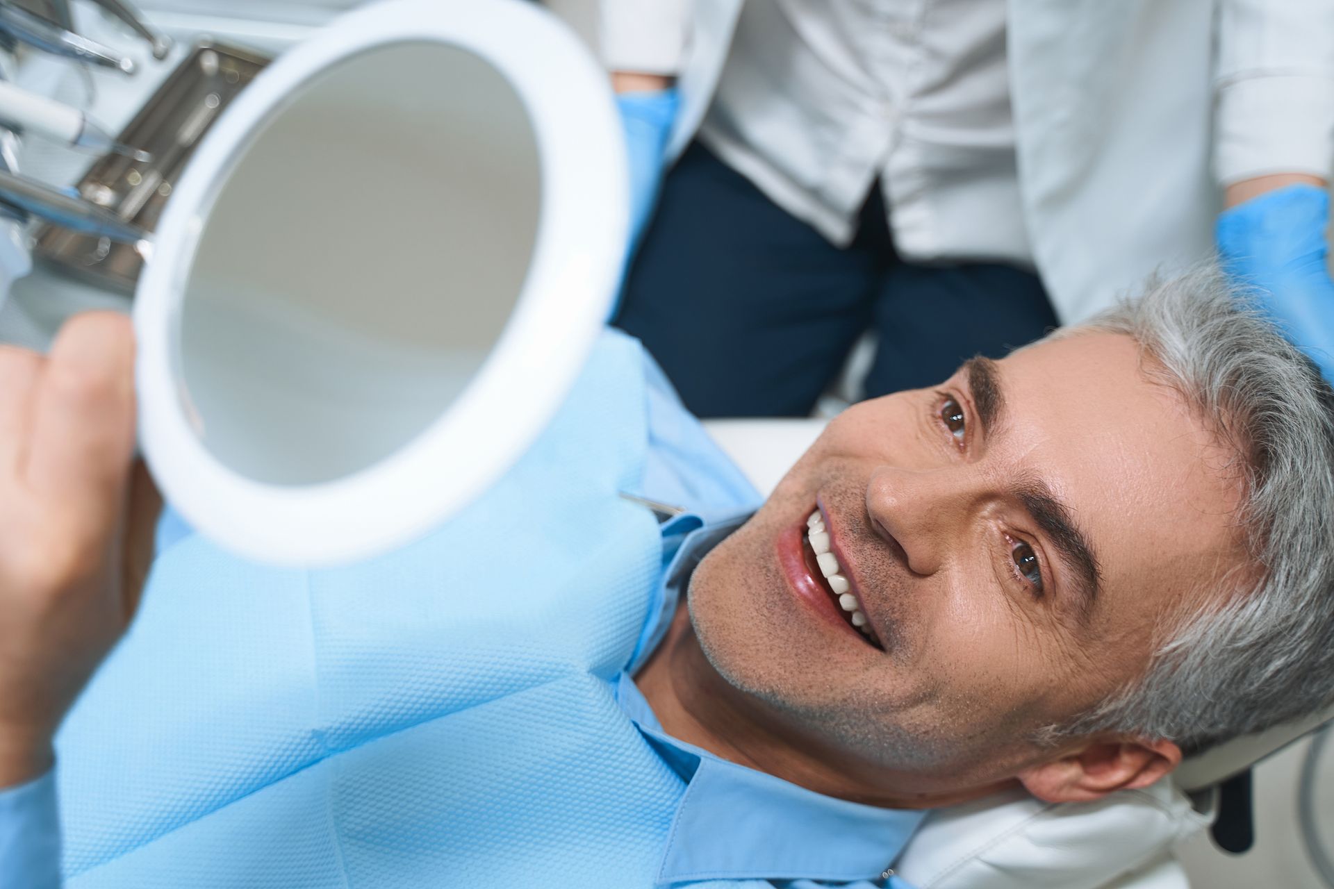 A man is sitting in a dental chair looking at his teeth in a mirror.