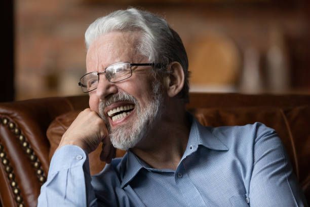 An older man with glasses is sitting on a couch and smiling.