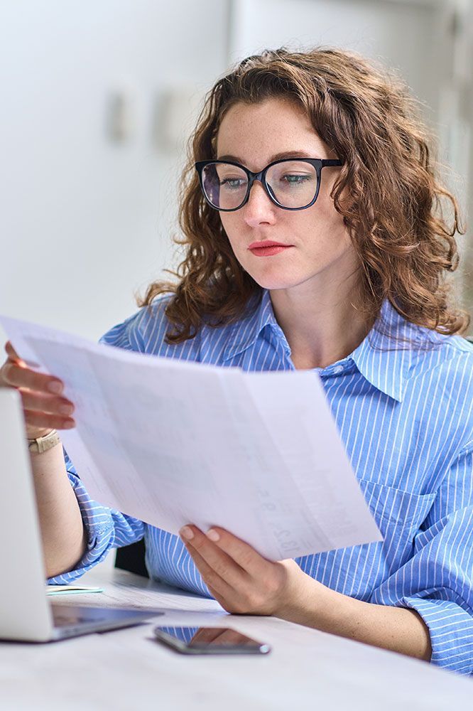 Woman with curly hair and glasses, looking at documents. She's in a blue shirt, next to a laptop and phone.
