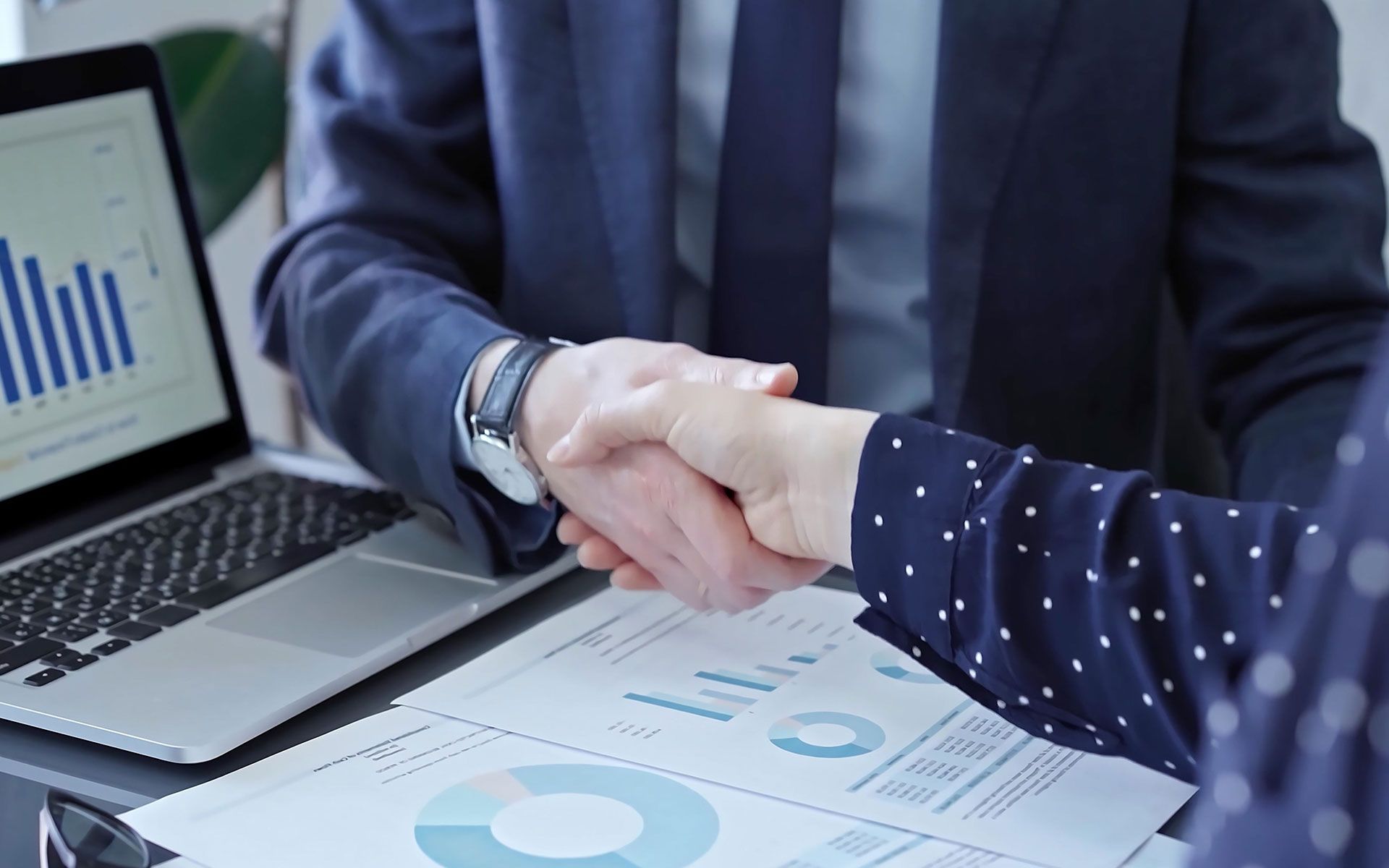 Two people shaking hands over documents and a laptop with a graph on the screen.