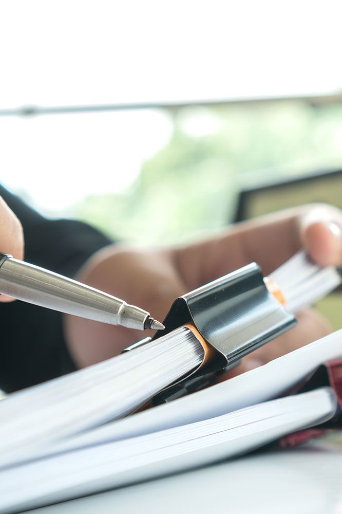 Person's hand with pen pointing to documents held in a binder clip.