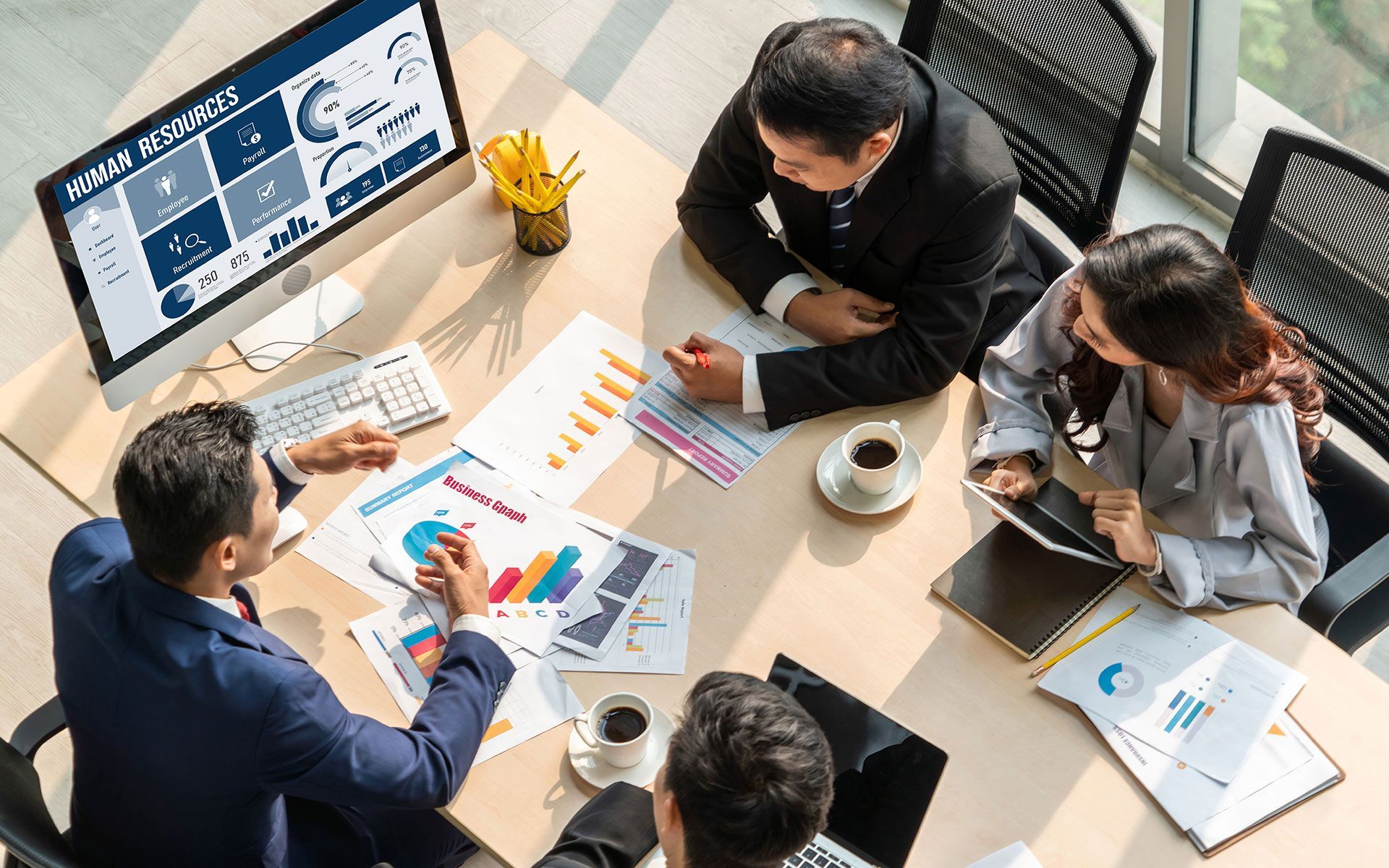 Business people at a table reviewing data charts and a computer screen in a bright office.