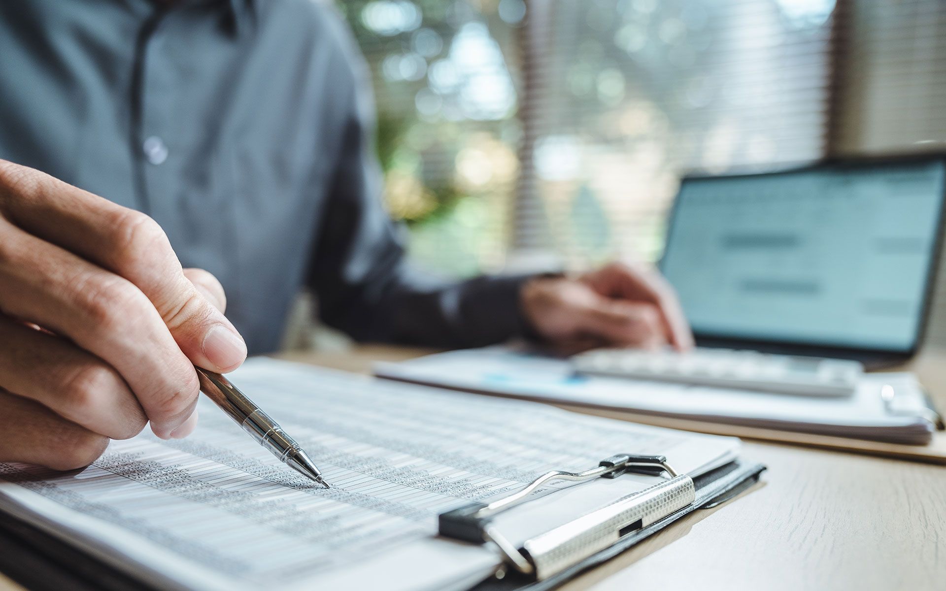 Person at desk, writing on a clipboard with a pen, laptop and calculator nearby.