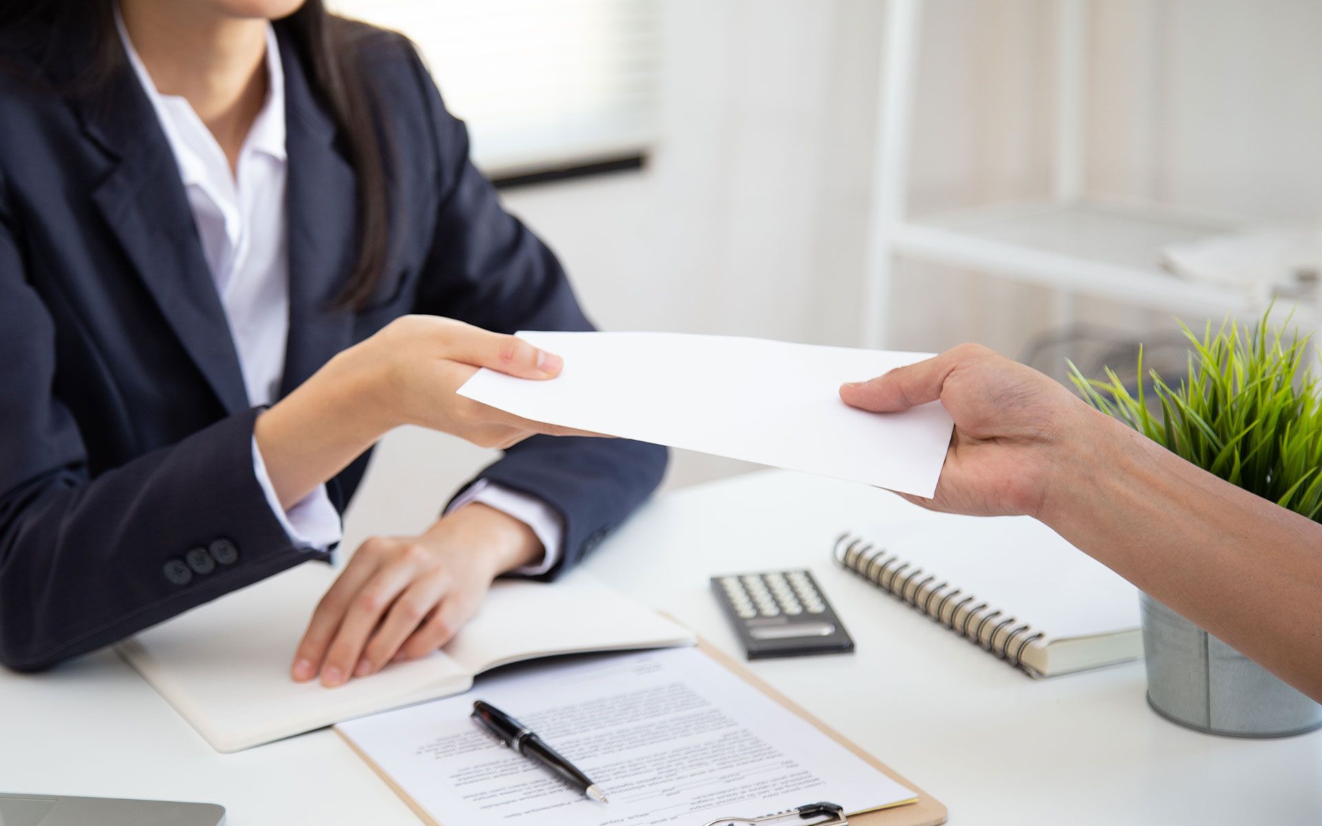 Person in blazer handing a document to another person at a desk.
