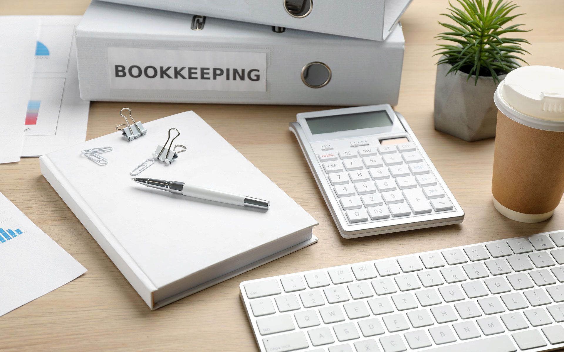 Bookkeeping office supplies including binders, calculator, keyboard, and notepad on a wooden desk.