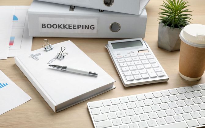 Bookkeeping office supplies including binders, calculator, keyboard, and notepad on a wooden desk.