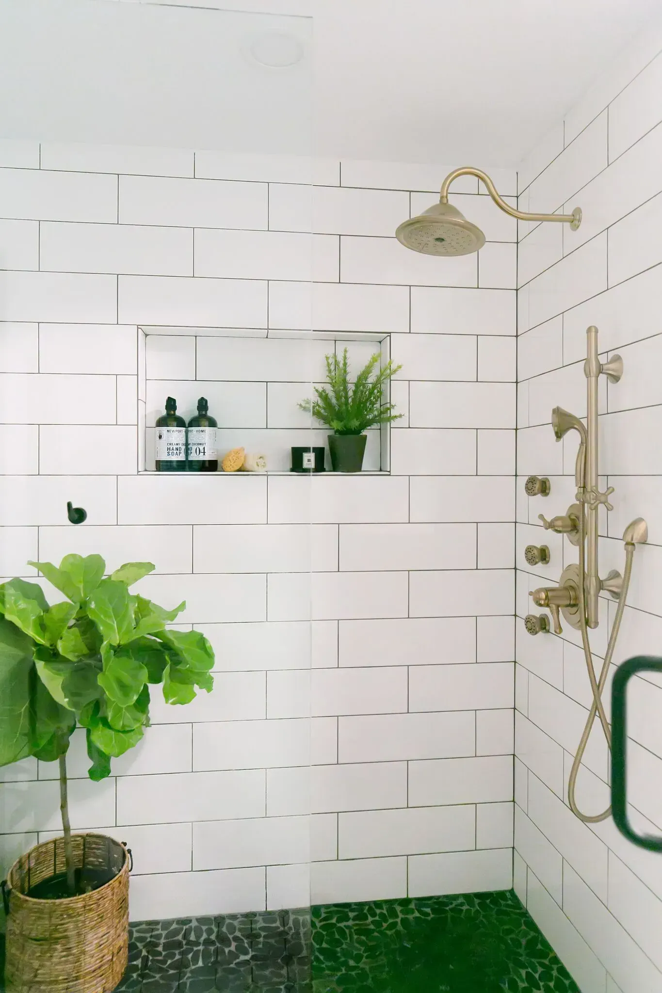A bathroom with white tiles , a shower , a plant and a shelf.