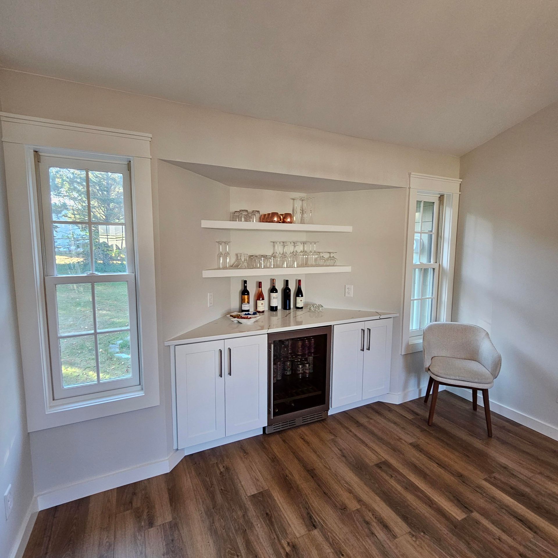 A living room with hardwood floors , white cabinets , a chair and a wine cooler.