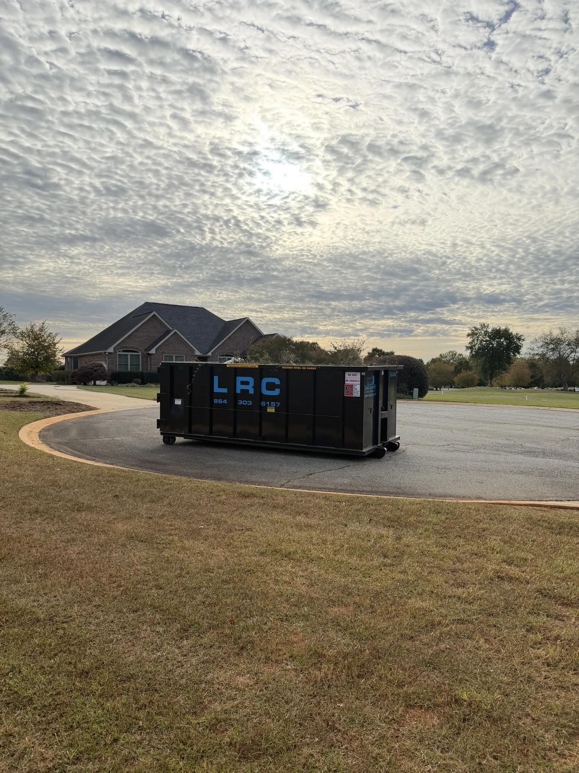 Black dumpster on a gravel driveway in front of a house, under a cloudy sky.