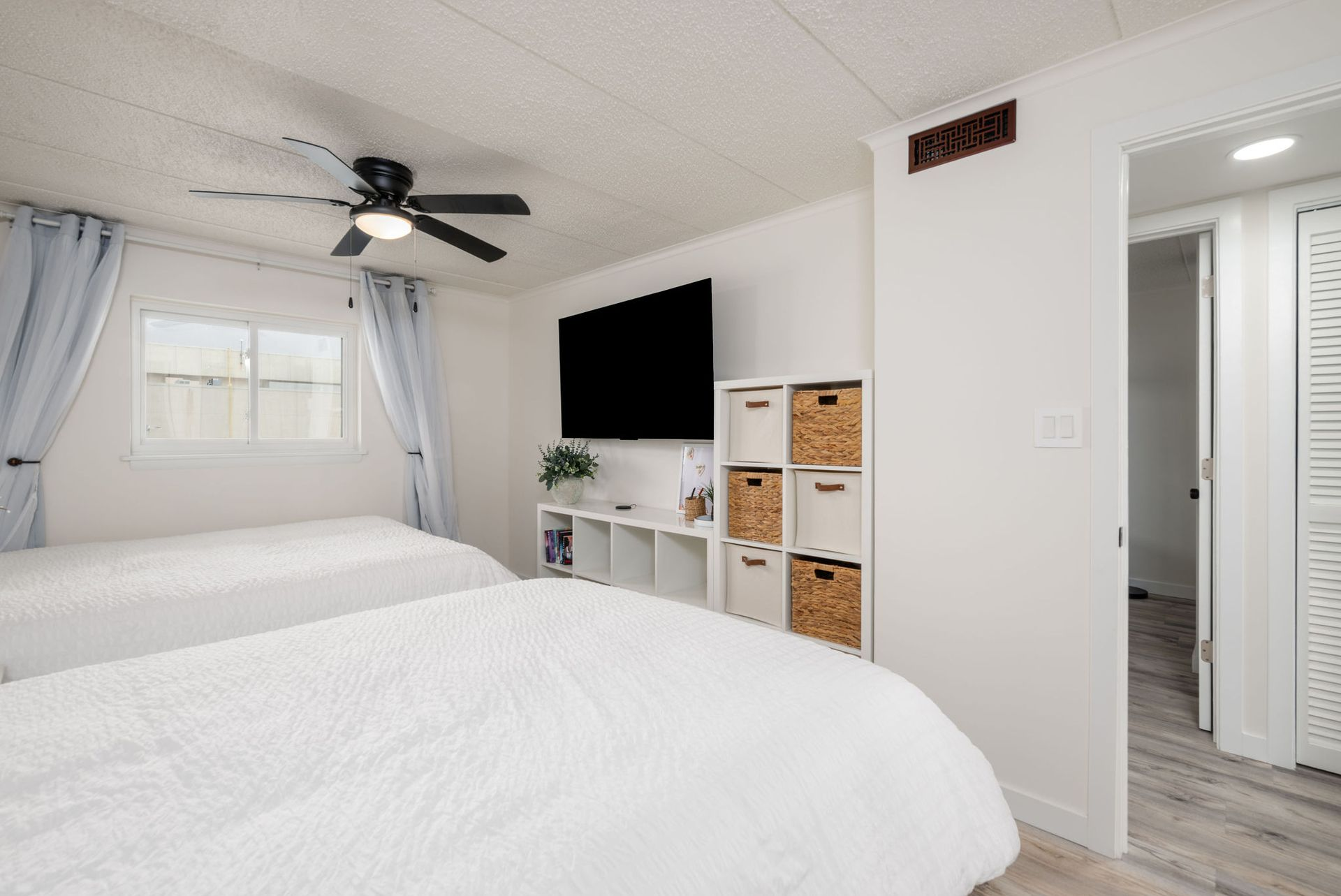 Bedroom with two white beds, a TV, and a cube shelf with baskets. Blue curtains and a ceiling fan.