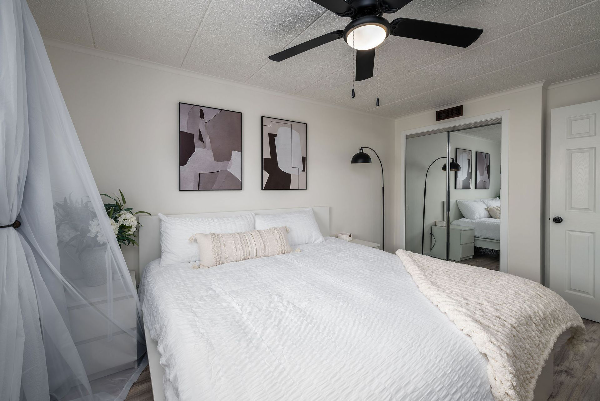 Bedroom with white bedding, abstract art, canopy, ceiling fan, and mirrored closet.