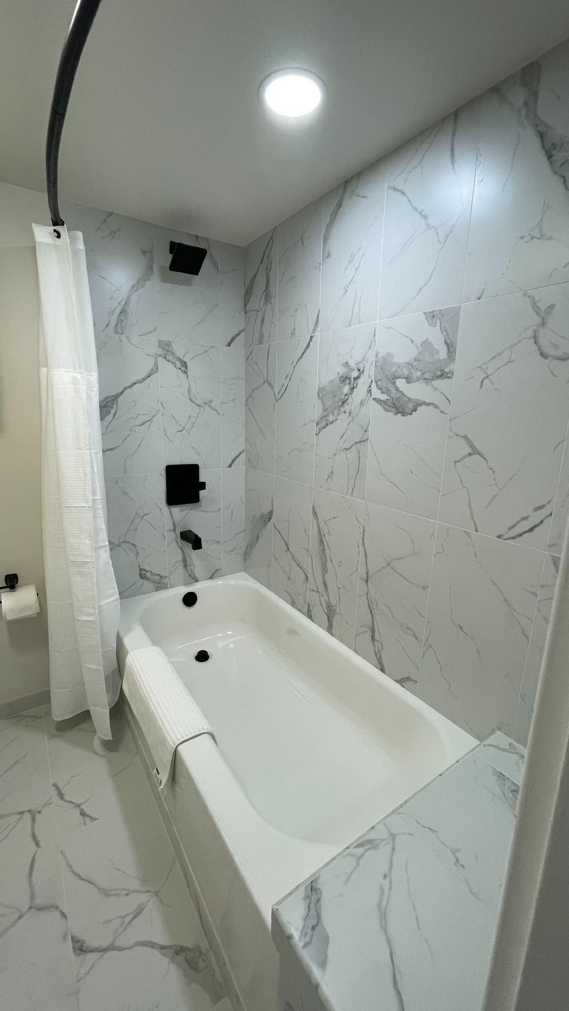 Bathroom with white marble-look tile, black fixtures, and a white bathtub with a shower curtain.