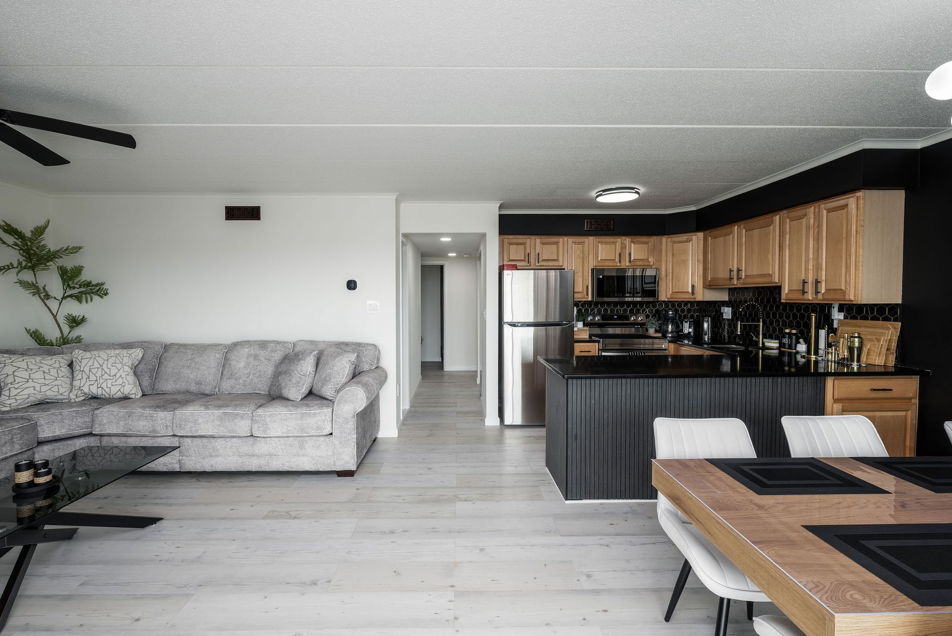 Open-concept living space with a kitchen, living area, and dining table. Gray sofa, light wood floors, and black accents.