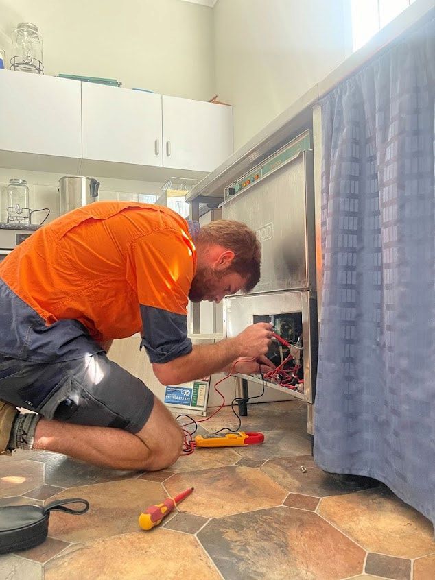 A Man In An Orange Shirt Kneels, Working On A Machine — Mackies in Taree, NSW