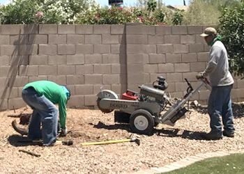 Two men are working on a stump grinder in front of a brick wall.