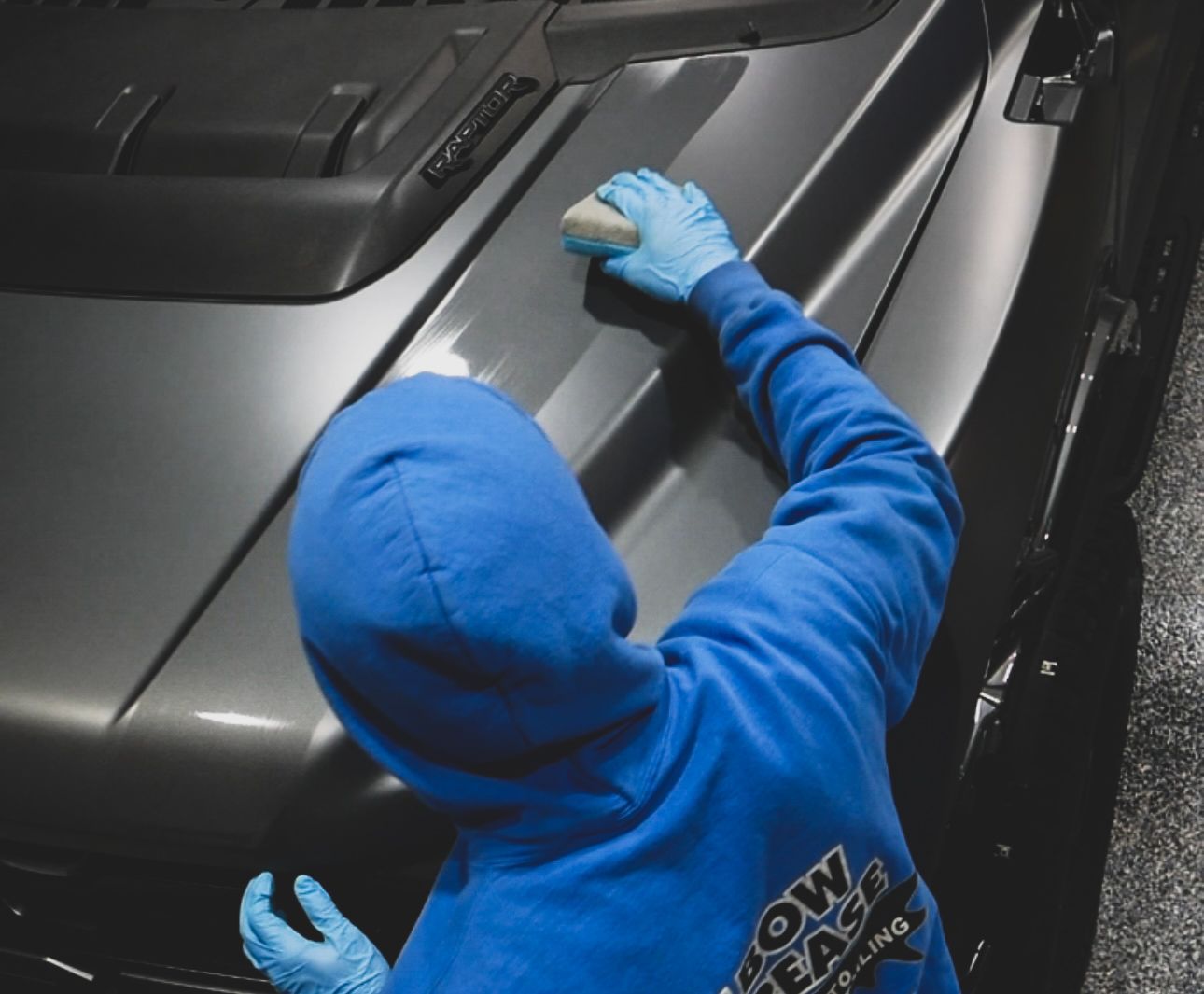 Person in a blue hoodie cleaning a gray car hood with a cloth.