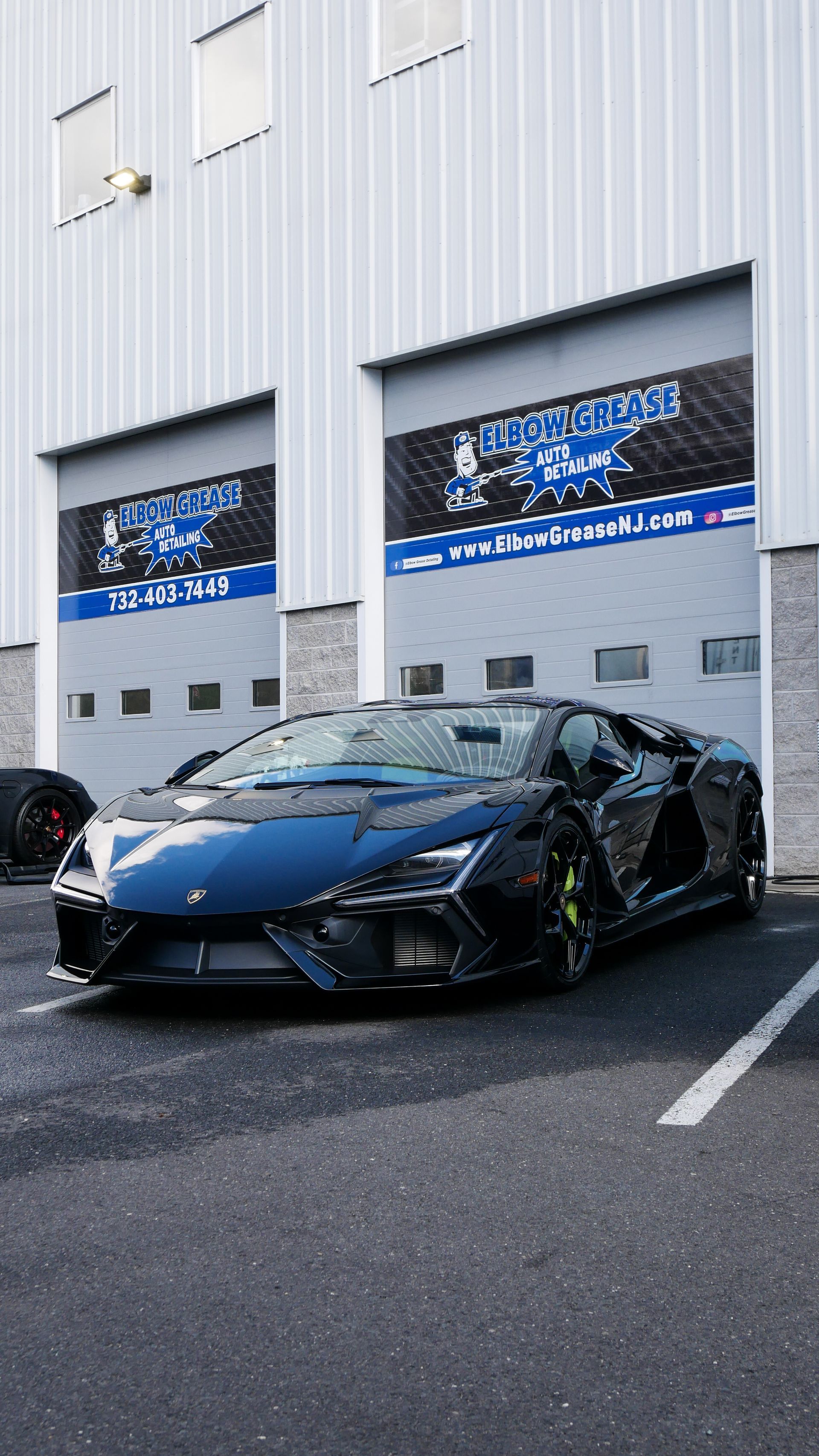 Dark blue Lamborghini Centenario parked in front of a garage with open doors and a company sign.