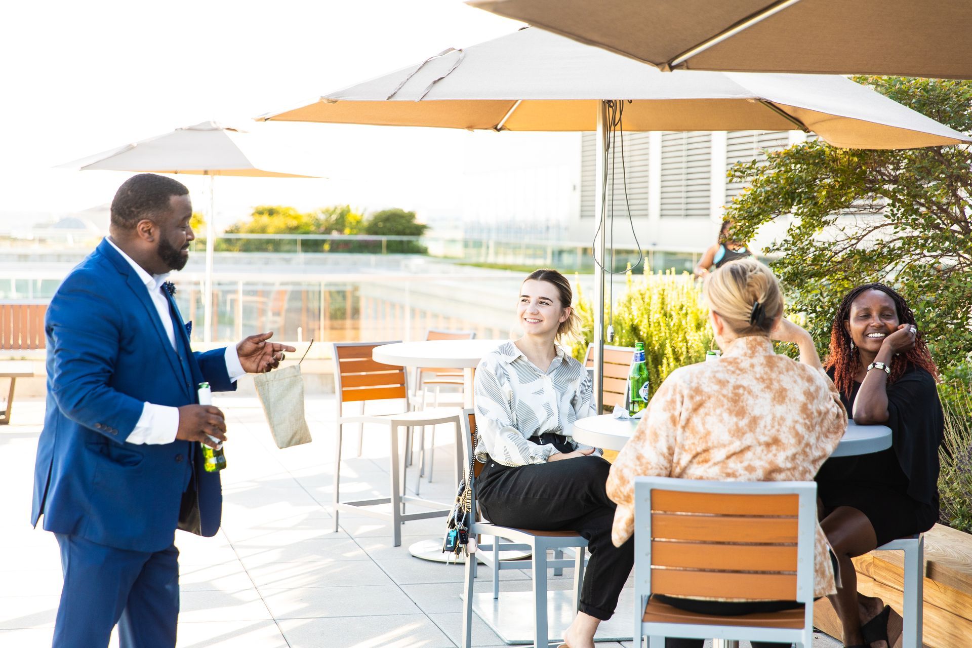 A man in a blue suit is standing next to a group of people sitting at tables under umbrellas.