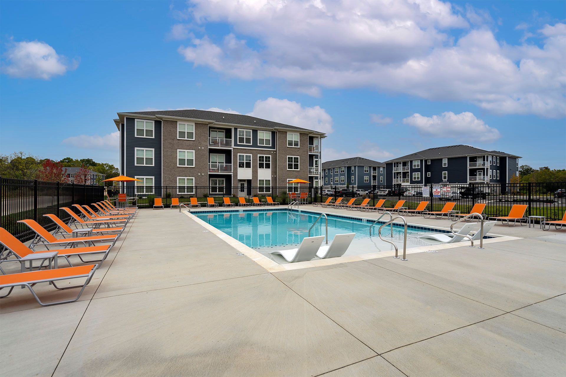 Outdoor apartment community pool with orange lounge chairs and surrounding buildings.