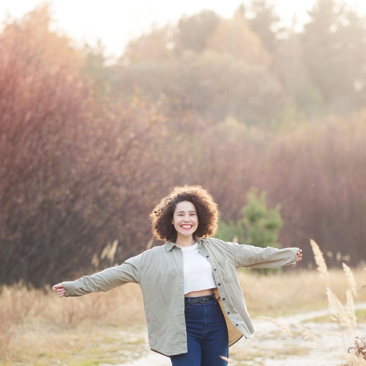 A woman with curly hair is standing in a field with her arms outstretched.