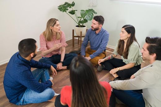 A group of people are sitting in a circle on the floor talking to each other.