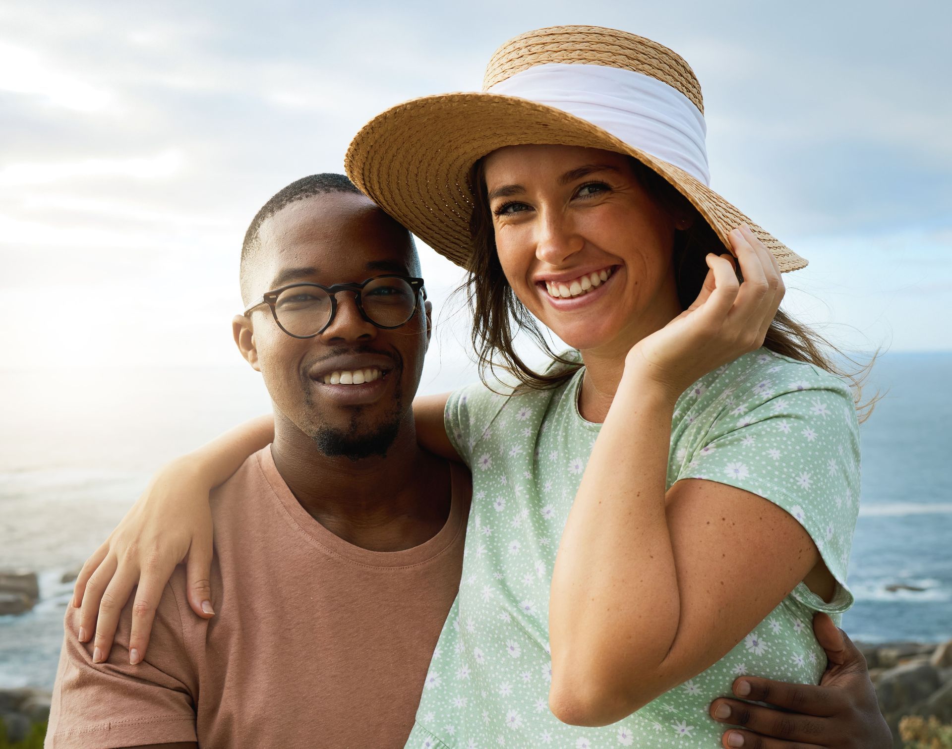 A happy man and a woman are posing for a picture in front of the ocean. The woman is wearing a straw hat.