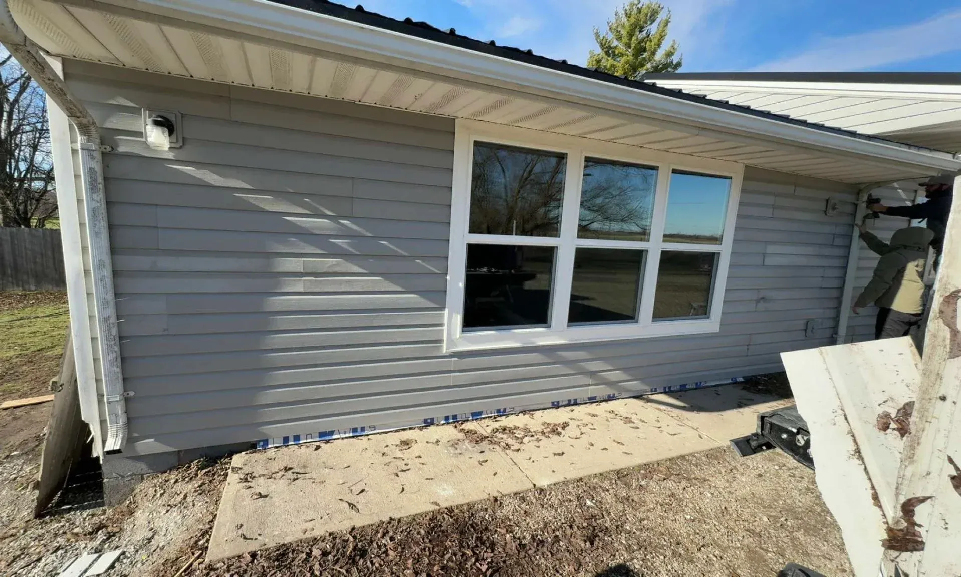 Exterior view of a house with gray siding and a large window. A person is partially visible on the right.