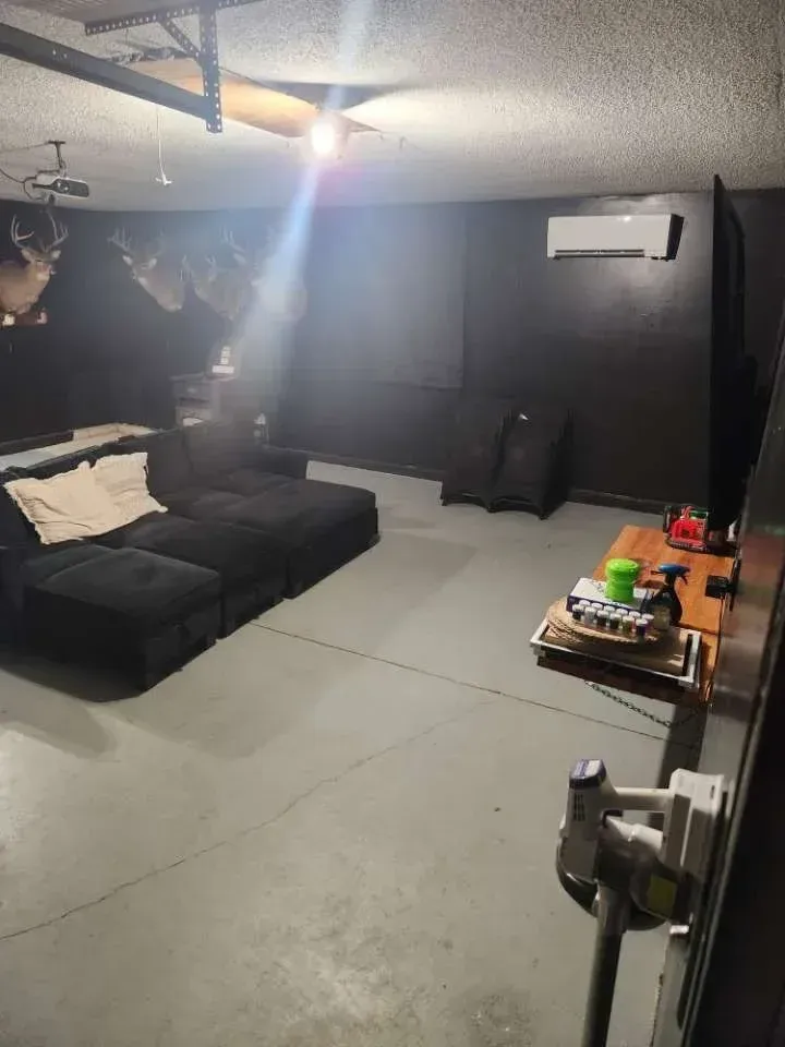 Dark-painted garage converted into a lounge with black couches and a grey floor. A/C unit and small table present.