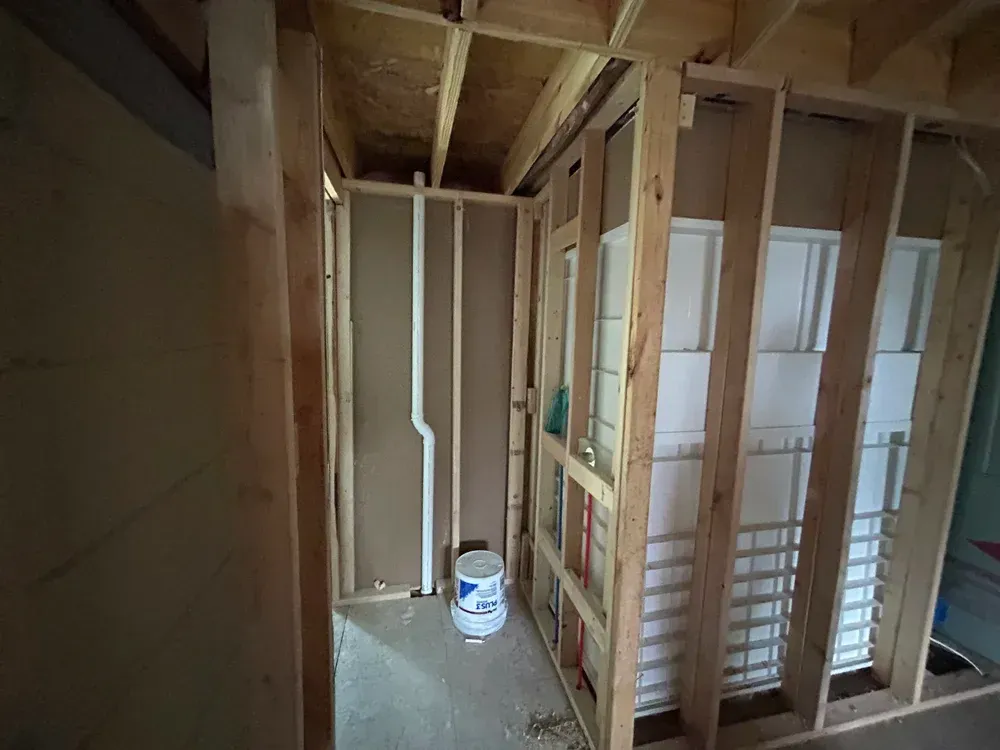 Interior view of a room under construction, wooden frames, plumbing visible, and a bucket on the floor.