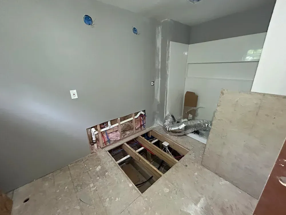 Bathroom under construction with exposed pipes and subfloor; gray walls.