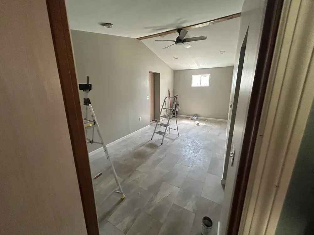 A room under renovation with grey walls, tile floor, a ladder, and a ceiling fan.