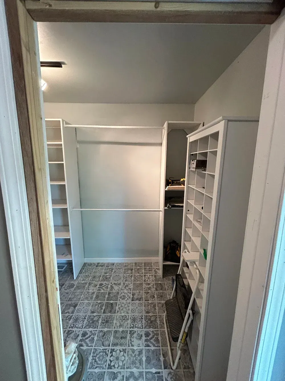 Walk-in closet with white shelves, hanging rods, and patterned floor seen through an unfinished doorway.