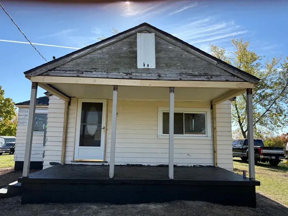 A small, weathered, yellow house with a front porch. Blue sky in the background.