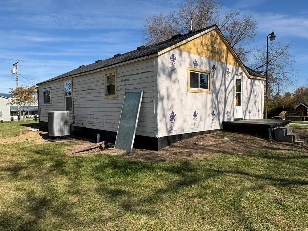House under construction; siding partially installed. Blue tarp, windows, doorway on the side. Outdoors.