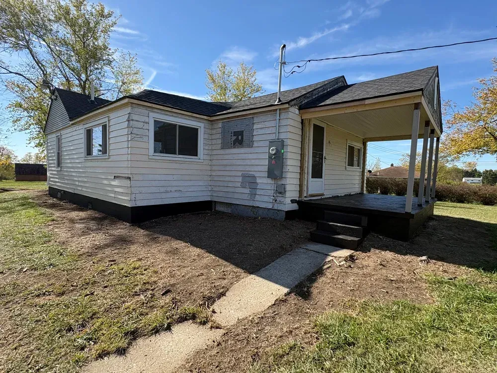 Small, white, one-story house with covered porch and black roof; situated in a grassy, rural area with blue sky.