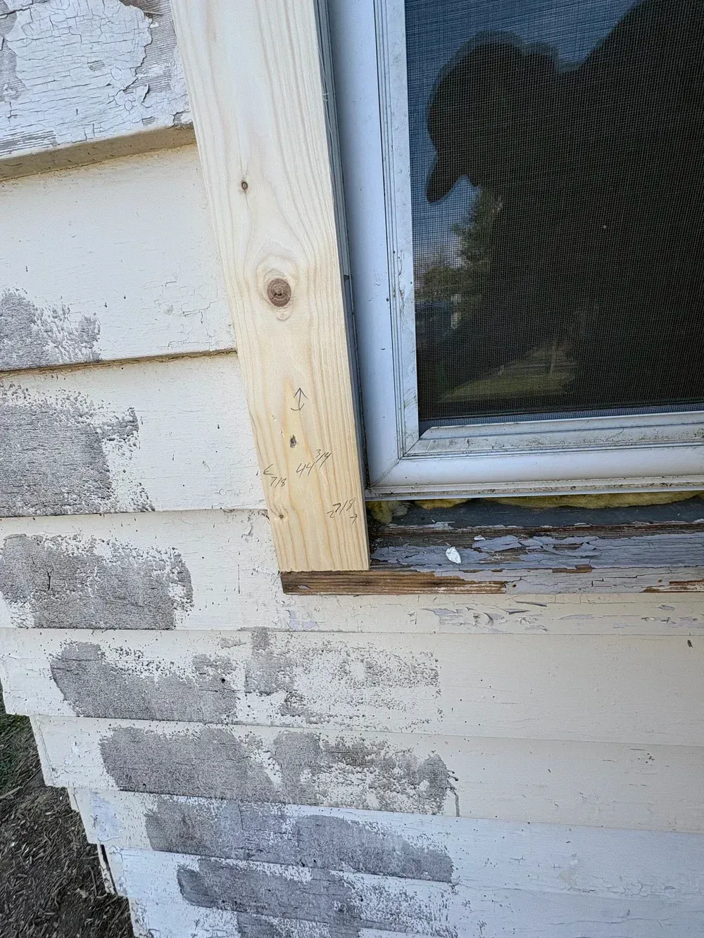 New wooden board next to a window, attached to white, peeling siding.