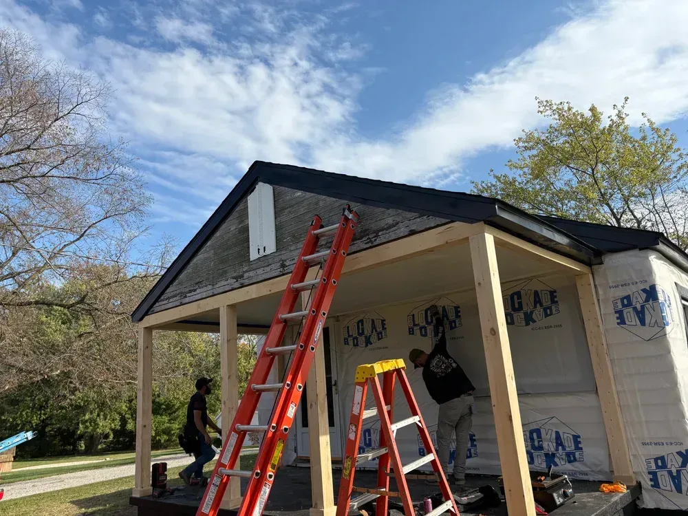 Construction workers building a porch on a small, white-sided structure with a black roof. Ladders and wood frames are visible.