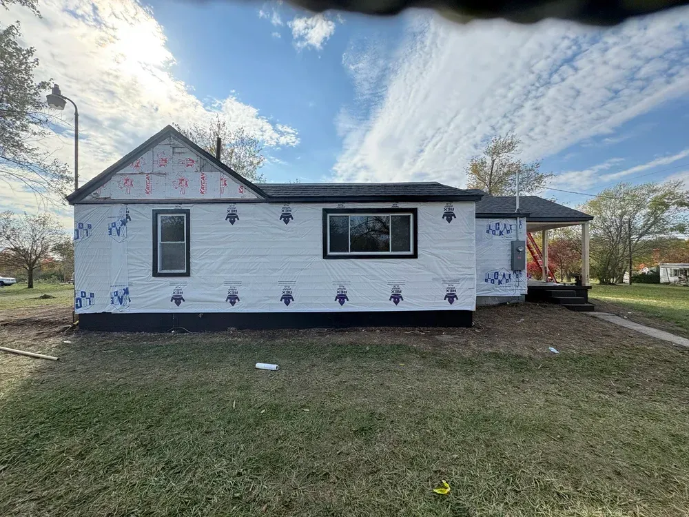 House under construction, white siding, black trim, blue wrap, overcast sky.