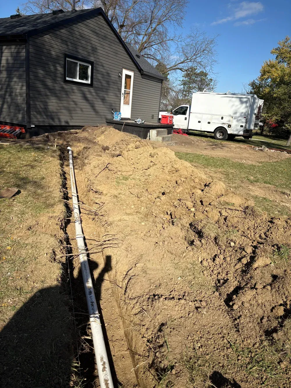 A trench with white pipes running towards a house, next to a dirt mound and a white truck.