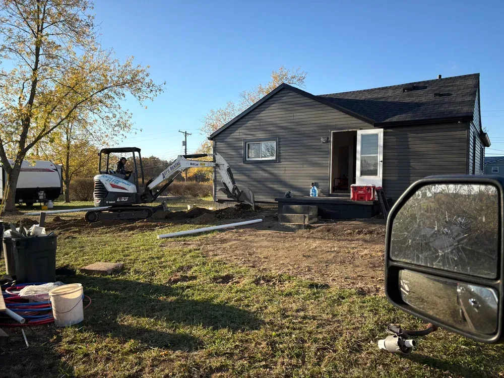 Construction site: Excavator digging near a small gray house, tools and supplies visible. Blue sky.