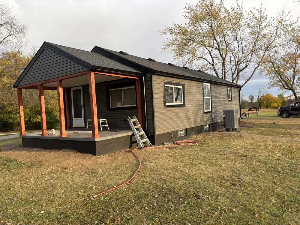 A gray-sided house with a porch and brown beams, set in a yard with dry grass.