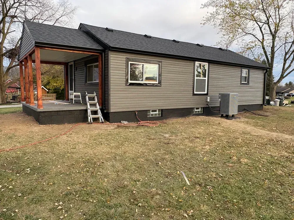 A gray-sided house with a porch and brown beams, set in a yard with dry grass.