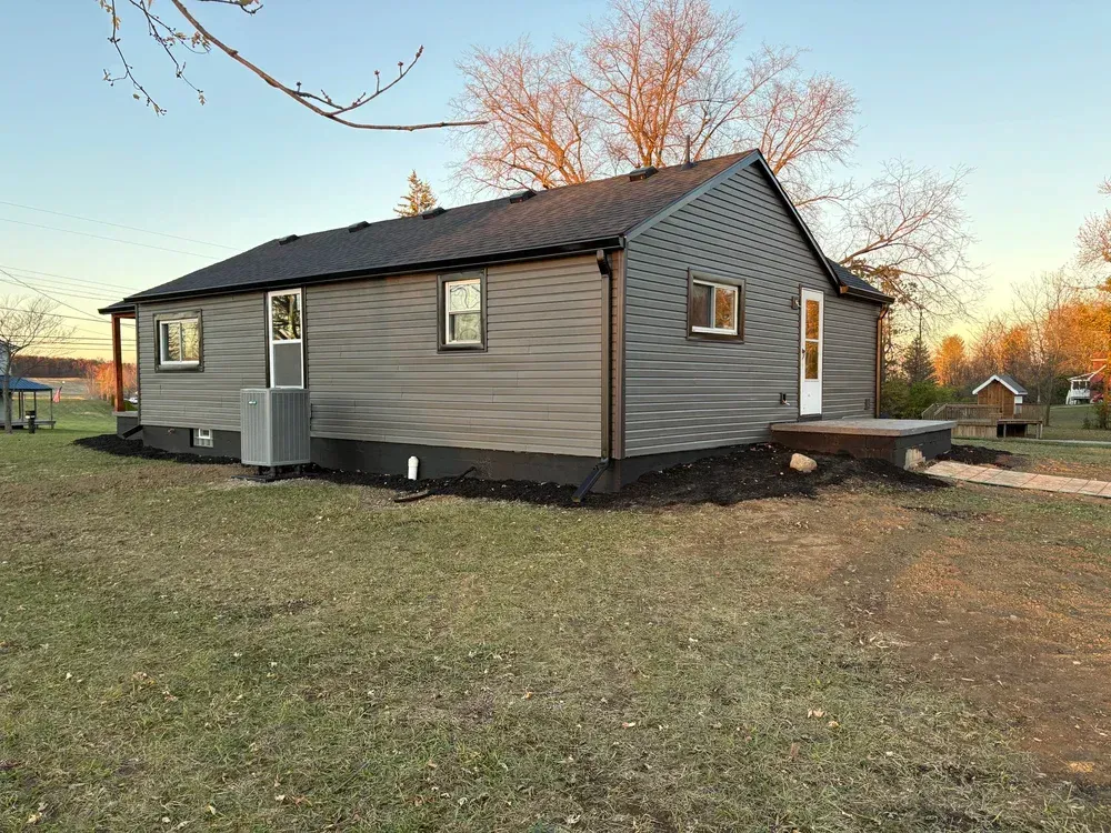 Gray-sided house with black trim and roof, surrounded by grass and mulch beds, set in a rural area.