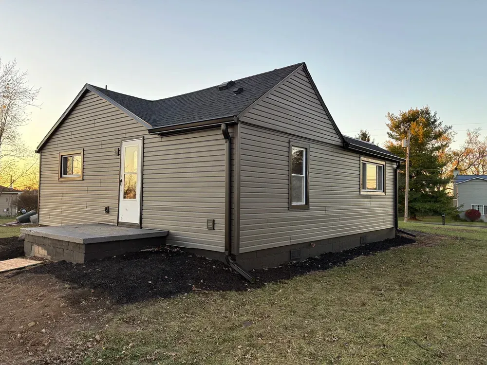 A gray house with a black roof and trim, a small front porch, and a dark mulched bed, in a grassy yard.