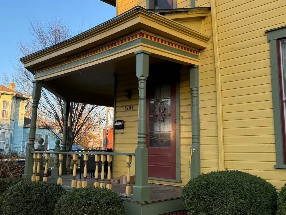 Yellow house with porch; red door, green trim, mailbox, and shrubbery.