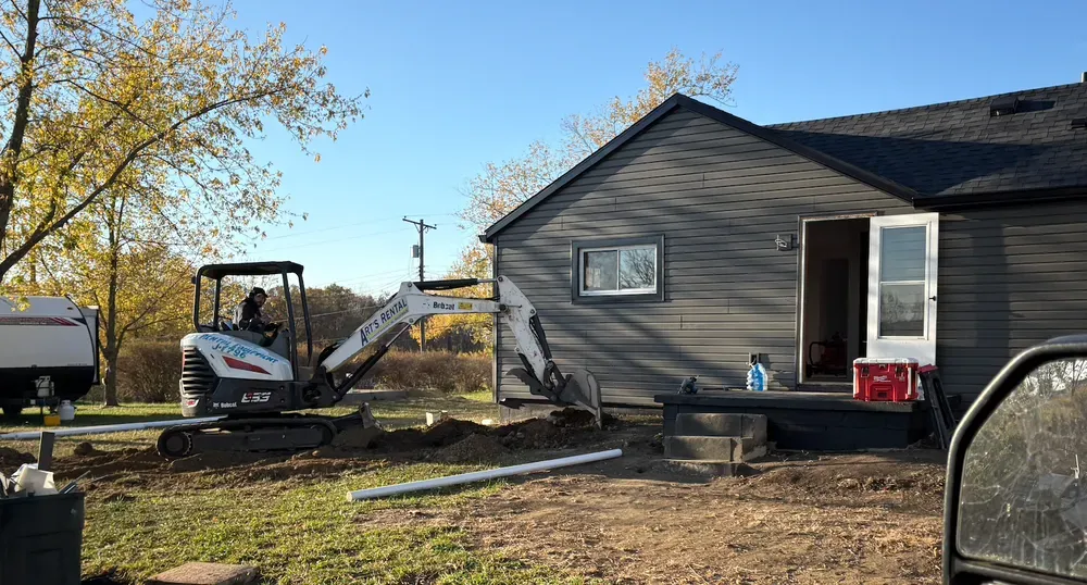A Bobcat excavator digging near a dark gray house with an open door, outdoors.