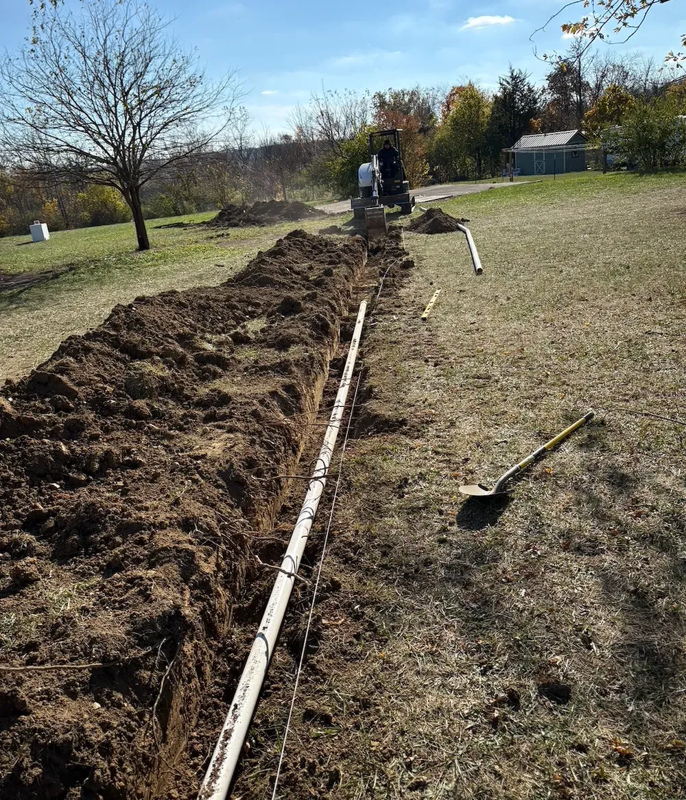 Trench dug in a grassy yard with white pipe inside; people working in background.