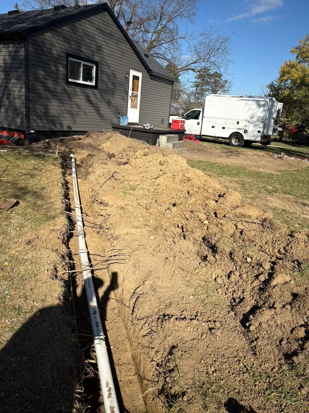 A trench dug in a yard with white PVC pipe, leading to a house and work truck.