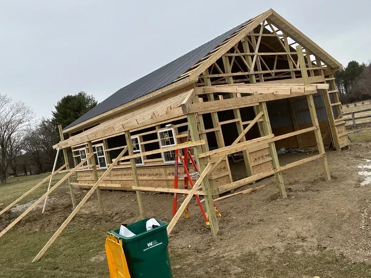 Construction of a wooden building with a partially installed roof, windows, and a porch. Green trash can is visible.