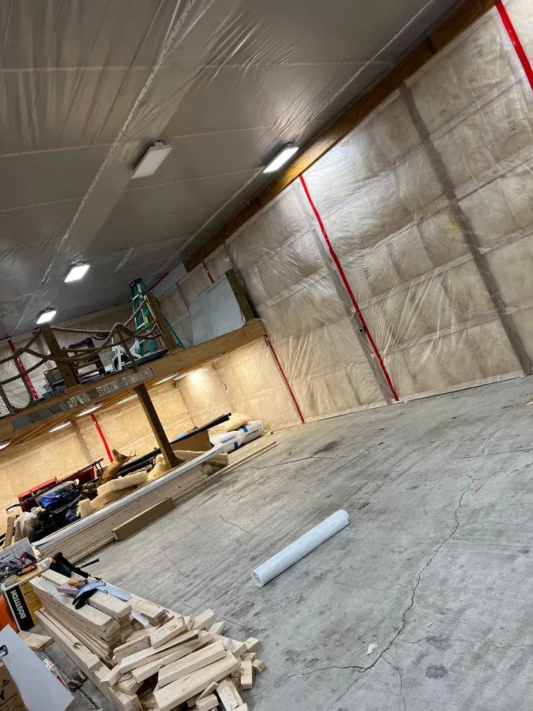 Construction in a large, unfinished warehouse. Walls have insulation covered in plastic; lumber and tools are scattered.